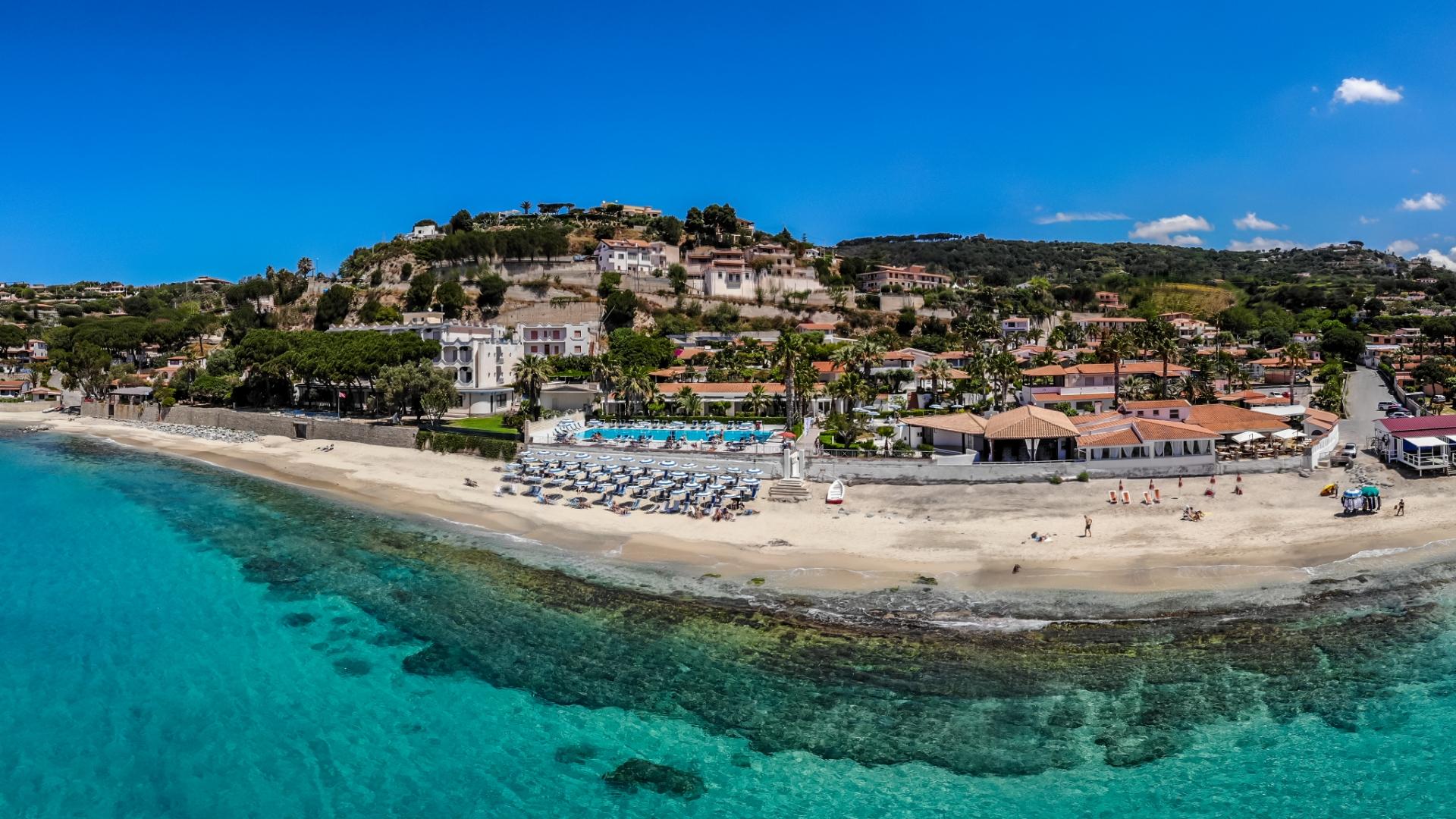 Spiaggia con mare cristallino, ombrelloni, e case collinari immerse nel verde.