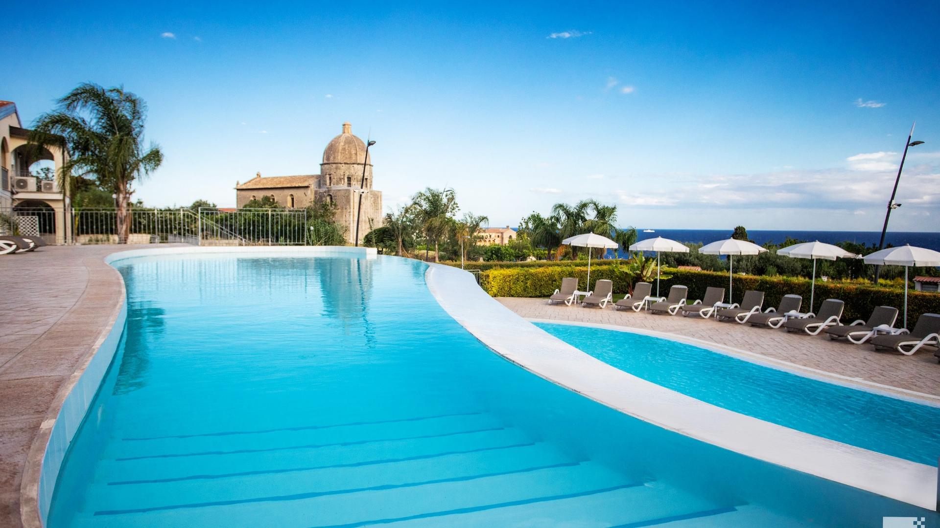 Piscina con vista mare, ombrelloni e edificio storico sullo sfondo.