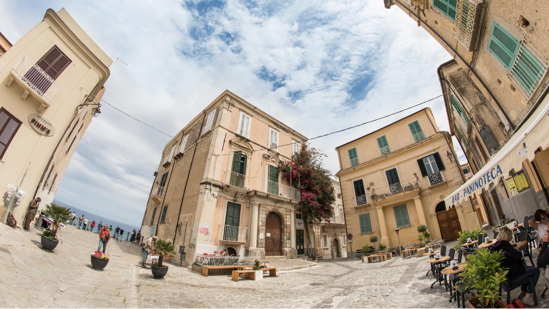 Piazza pittoresca con edifici storici e caffè all'aperto, vista sul mare.