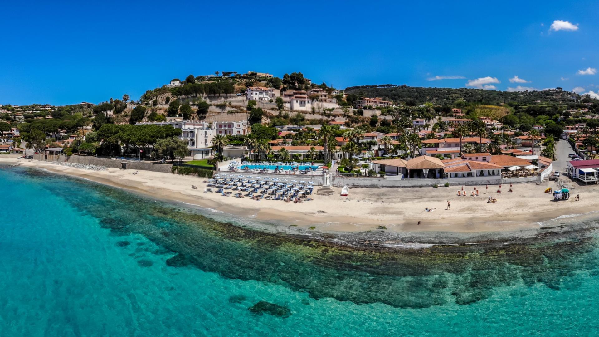 Spiaggia con ombrelloni, mare cristallino e case collinari sotto un cielo azzurro.