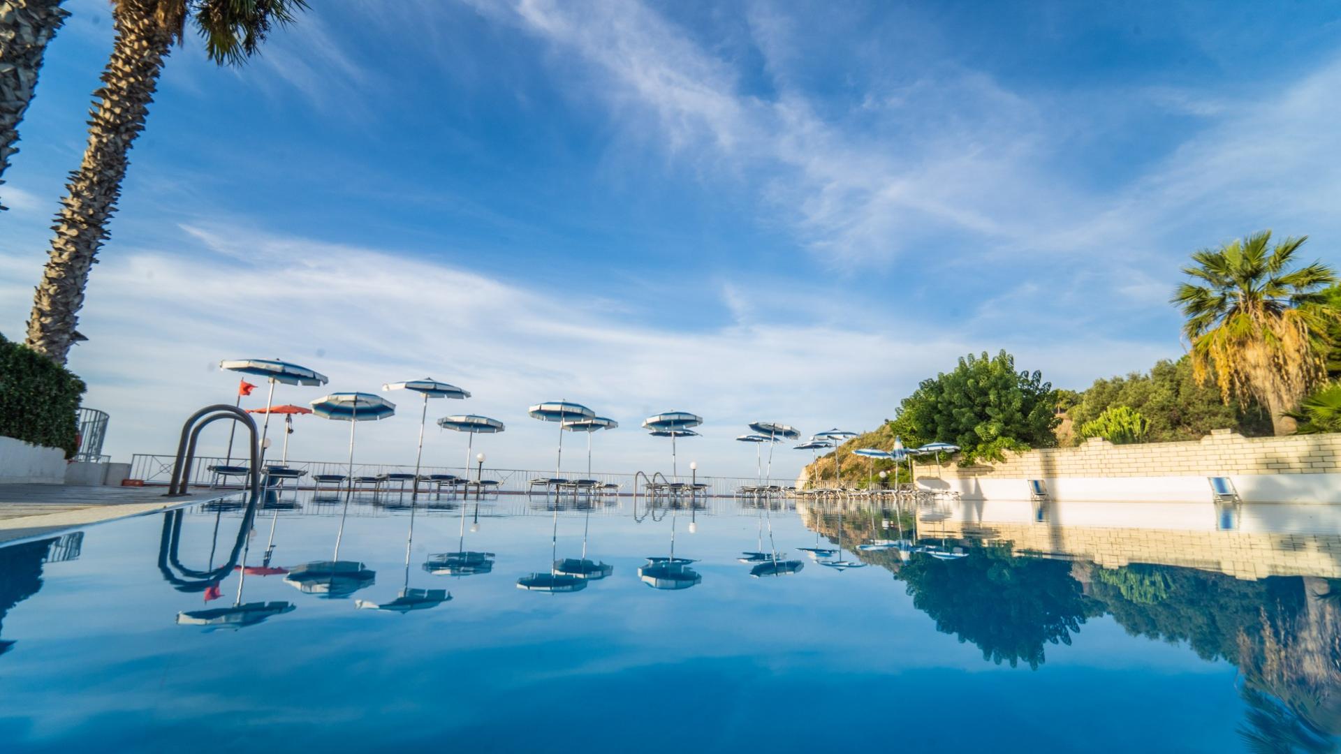 Piscina all'aperto con ombrelloni e cielo sereno riflessi nell'acqua cristallina.