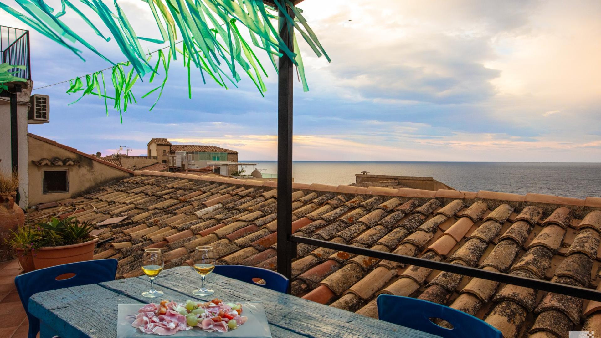 Terrazza con vista mare, tetti di tegole, due bicchieri di vino e antipasto.