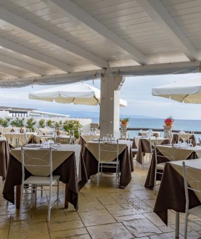 Ristorante all'aperto con vista mare, tavoli apparecchiati sotto ombrelloni bianchi.