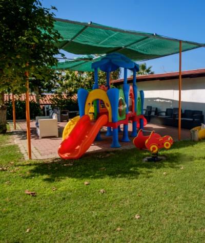 Area giochi per bambini all'aperto con scivolo e sedute ombreggiate.