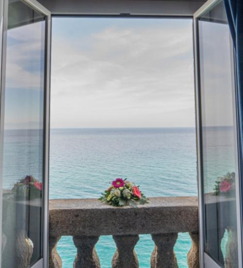 Finestra aperta sul mare con fiori sul balcone e tende blu.