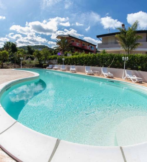 Piscina all'aperto con lettini, cielo sereno e case sullo sfondo.