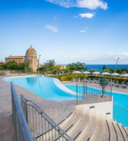 Piscina con vista mare, circondata da lettini e ombrelloni, vicino a una chiesa.