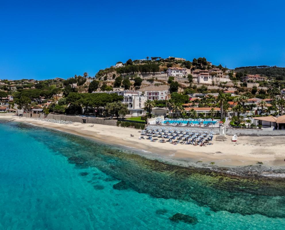 Spiaggia con mare cristallino, ombrelloni, e case collinari immerse nel verde.