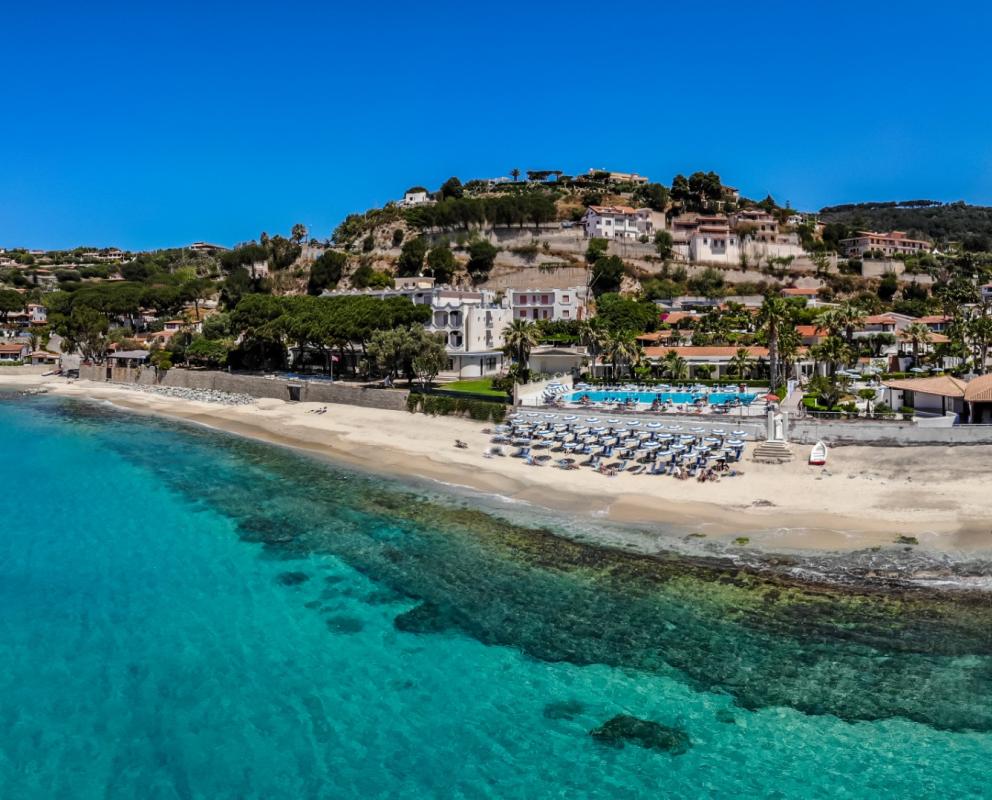 Spiaggia con ombrelloni, mare cristallino e case collinari sotto un cielo azzurro.