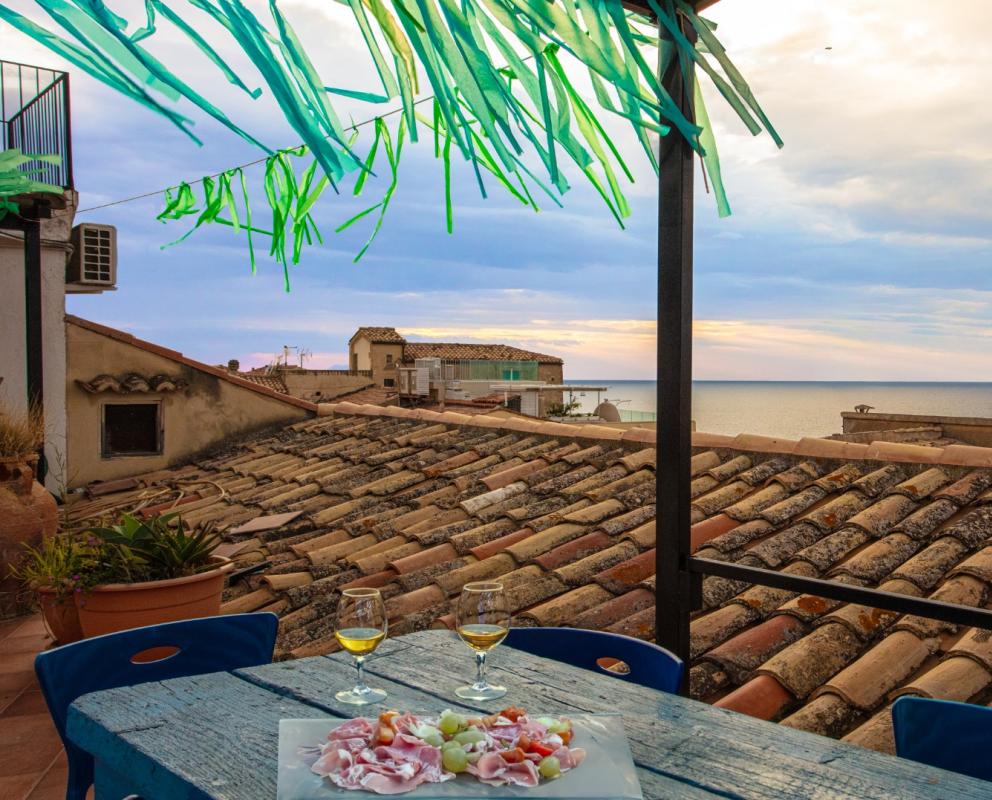 Terrazza con vista mare, tetti di tegole, due bicchieri di vino e antipasto.