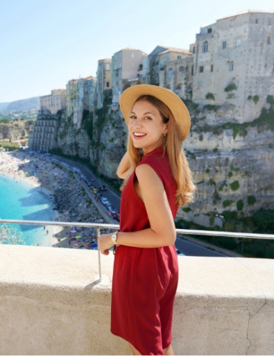 Donna sorridente in abito rosso e cappello, con vista su una città costiera.