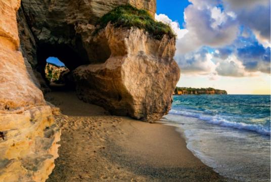 Spiaggia rocciosa con grotta, onde delicate e cielo nuvoloso al tramonto.