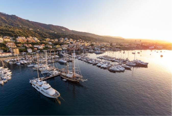 Porto turistico con yacht e barche a vela al tramonto, colline sullo sfondo.